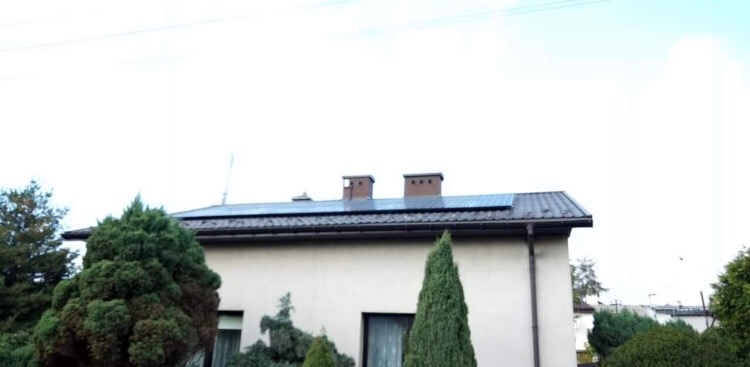 Dom jednorodzinny z panelami słonecznymi na dachu, widoczne dwa kominy i ozdobne iglaki w ogrodzie.