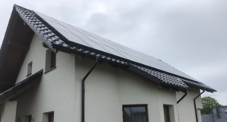 Panele słoneczne na dachu domu jednorodzinnego z ciemną dachówką i orynnowaniem, pochmurne niebo w tle.
