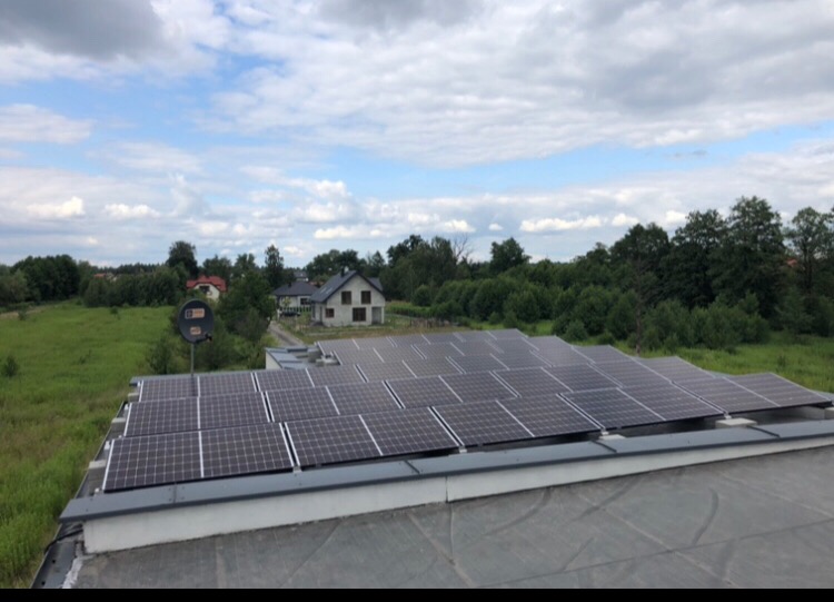 Instalacja paneli słonecznych na płaskim dachu budynku z widokiem na zielone tereny i domy w tle, uchwycona w pochmurny dzień.