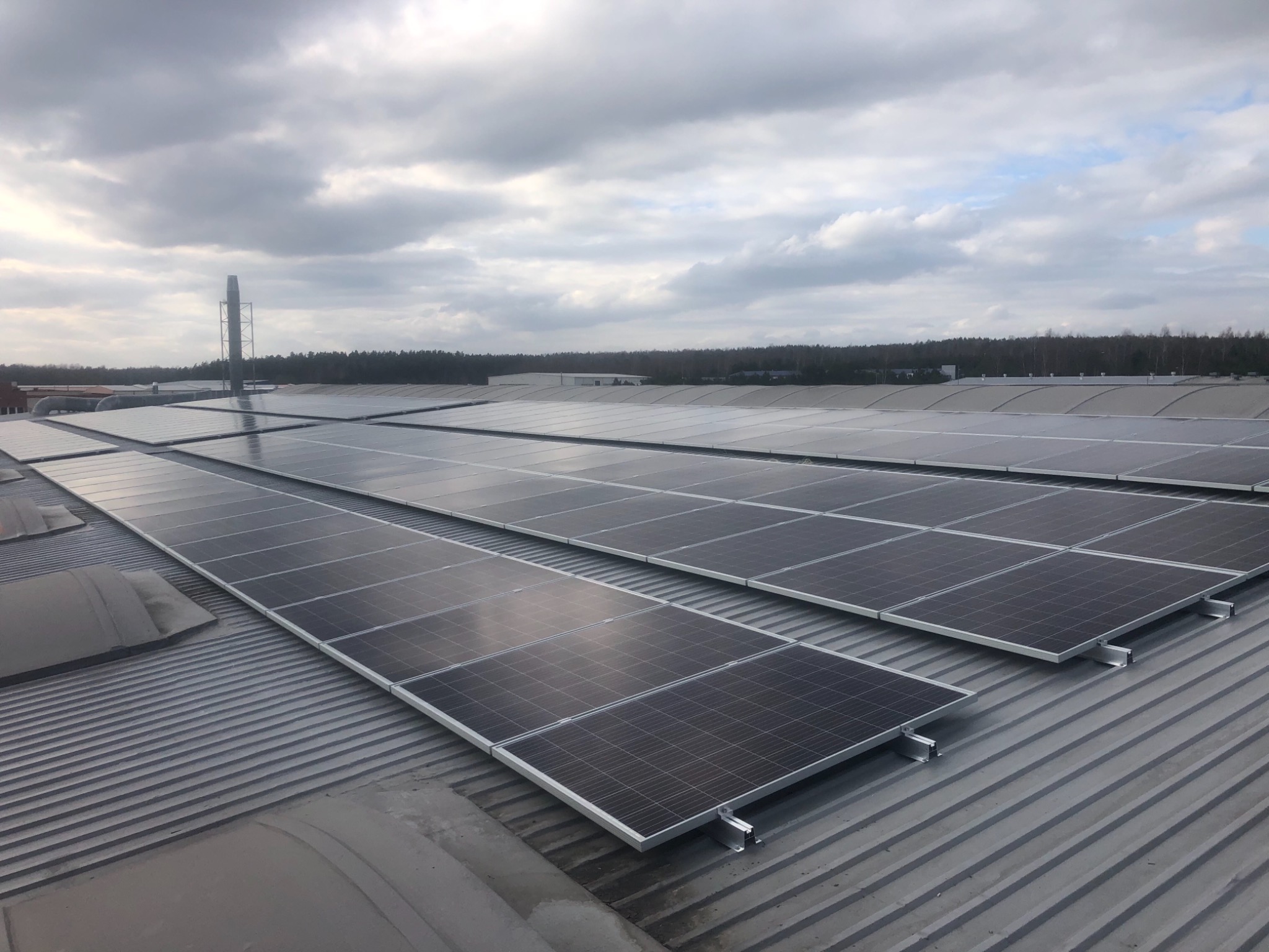 Instalacja fotowoltaiczna na dachu przemysłowym z widocznym kominem w tle, pochmurne niebo.