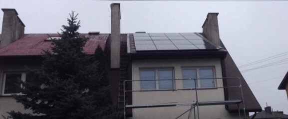 Instalacja paneli słonecznych na dachu dwupiętrowego domu, widoczne rusztowanie i zielone drzewo iglaste z przodu.