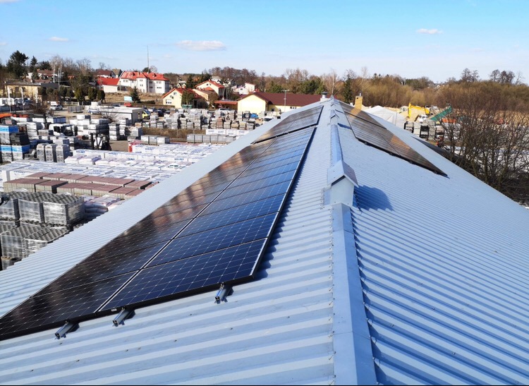 Panele słoneczne zamontowane na blaszanym dachu budynku przemysłowego, widok z góry, w tle zabudowa jednorodzinna i składowisko kostki brukowej.