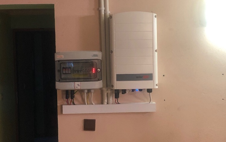 Zamontowany inwerter SolarEdge obok skrzynki elektrycznej KENO na ścianie w pomieszczeniu, widoczne kable i rurki instalacyjne.