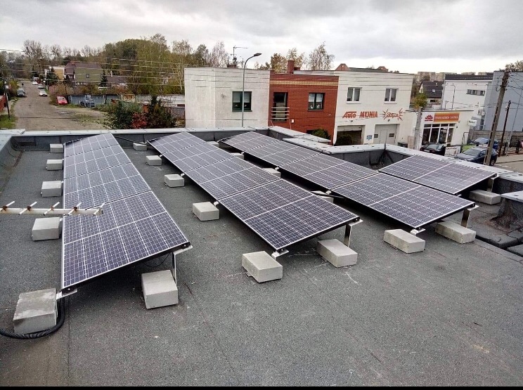 Panele słoneczne zamontowane na płaskim dachu budynku, wsparte na betonowych blokach, z anteną telewizyjną w tle.