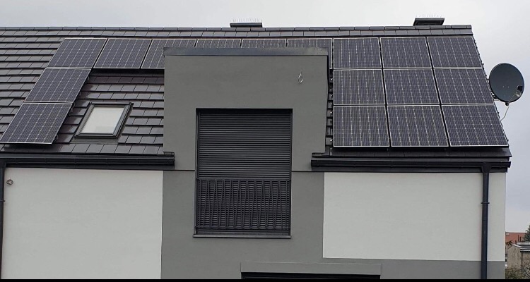 Panele słoneczne na dachu domu jednorodzinnego z brązową dachówką, widoczny również lukarna i antena satelitarna.