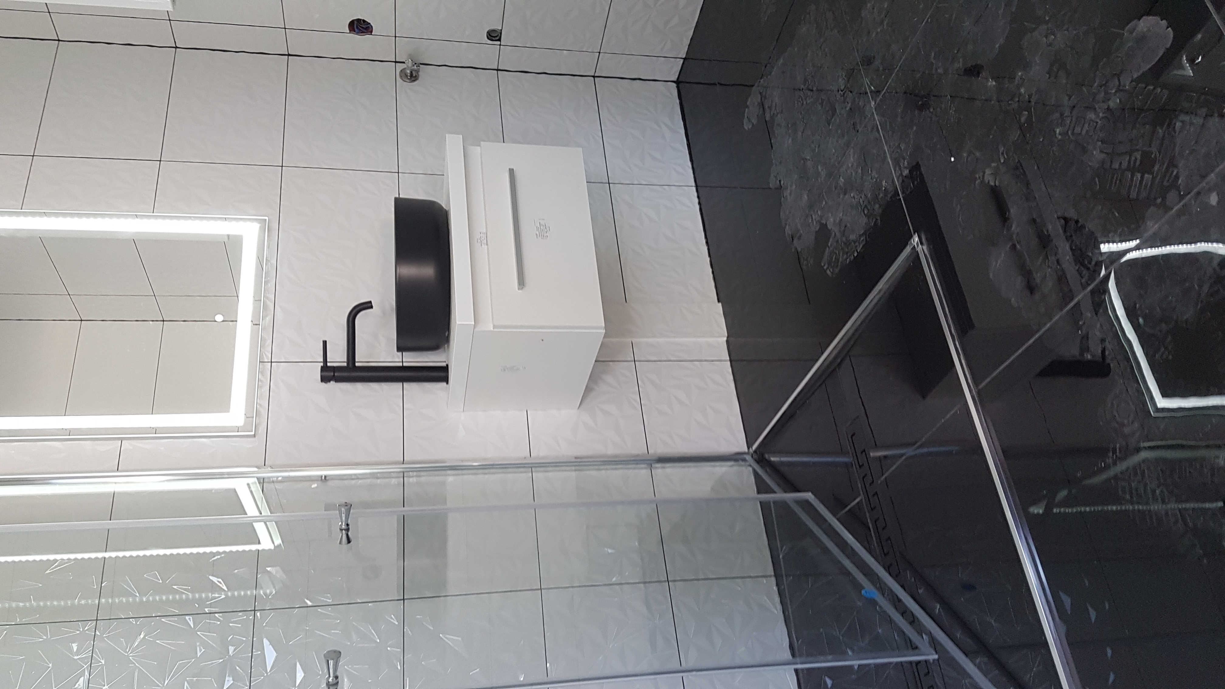 Nowoczesna łazienka z biało-czarną kolorystyką, kabiną prysznicową ze szklanymi drzwiami, umywalką nablatową w kolorze czarnym, białą szafką pod umywalką oraz lustrem z podświetleniem LED.