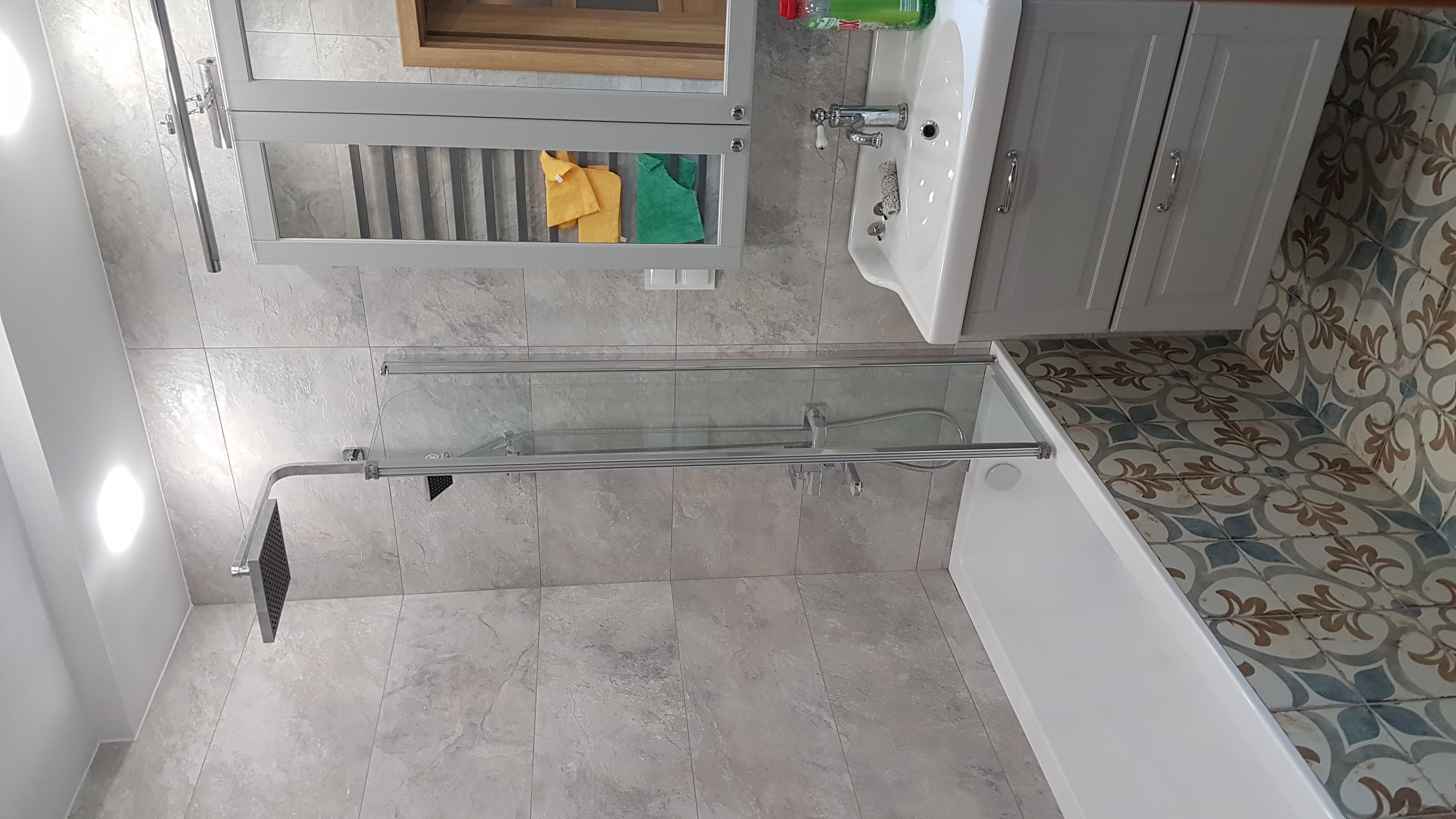 Nowoczesna łazienka z szarymi płytkami, kabiną prysznicową typu walk-in, białą umywalką z szafką i lustrem z półkami, oraz dekoracyjnymi płytkami podłogowymi.