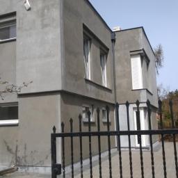 Usługi ogólnobudowlane - Szary, dwukondygnacyjny dom z płaskim dachem, widziany zza czarnego, kutego ogrodzenia. Widoczne okna z białymi ramami i fragment zielonego budynku w tle.