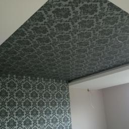 Usługi ogólnobudowlane - Pokój po remoncie: wzorzysta tapeta na ścianie i skośnym suficie, białe listwy przysufitowe, fragment okna. Wykończenie wnętrza w klasycznym stylu.
