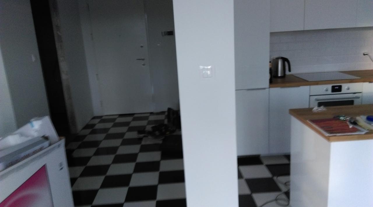Wnętrze mieszkania z czarno-białą szachownicą na podłodze, widoczna kuchnia z białymi szafkami i drewnianym blatem oraz fragment przedpokoju z drzwiami.
