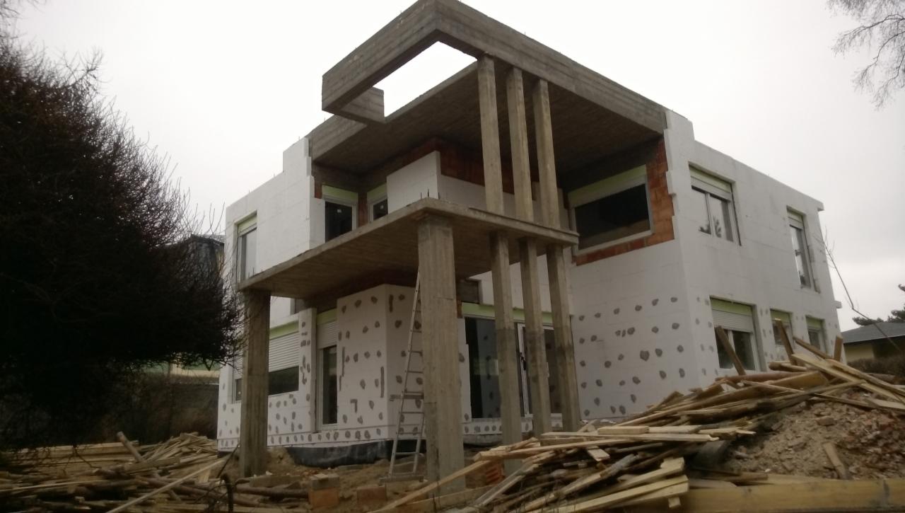 Budynek w trakcie ocieplania styropianem, widoczne betonowe elementy konstrukcyjne i rusztowanie. Na pierwszym planie leżące deski.