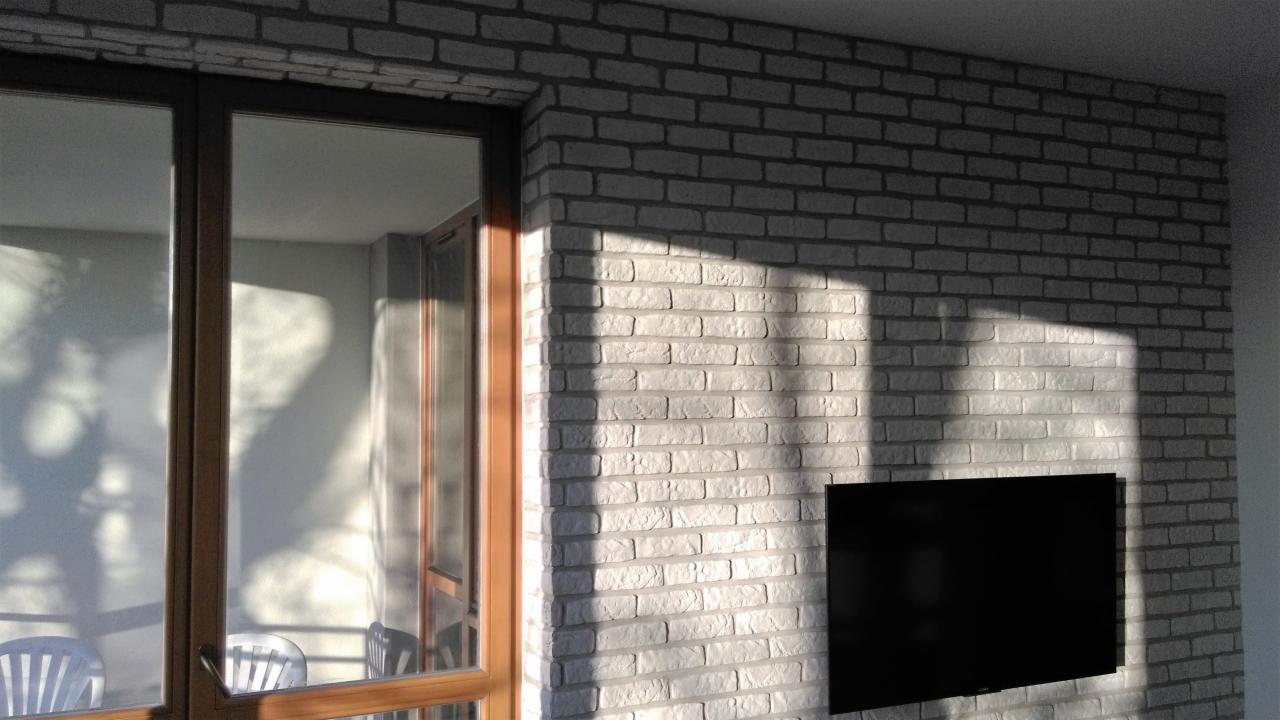 Biała ściana z cegły dekoracyjnej w salonie z oknem balkonowym i telewizorem. Fragment balkonu z krzesłami. Oświetlenie słoneczne.