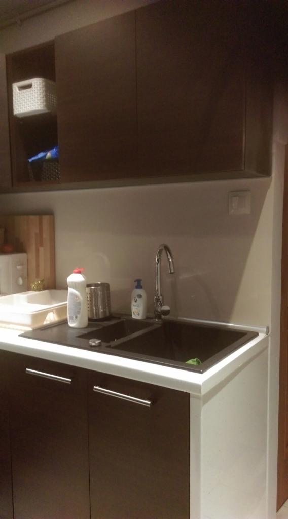 Ciemna zabudowa kuchenna z dwukomorowym zlewem, srebrną baterią i białym blatem. Nad zlewem wiszące szafki. Widoczne środki czystości i akcesoria kuchenne.