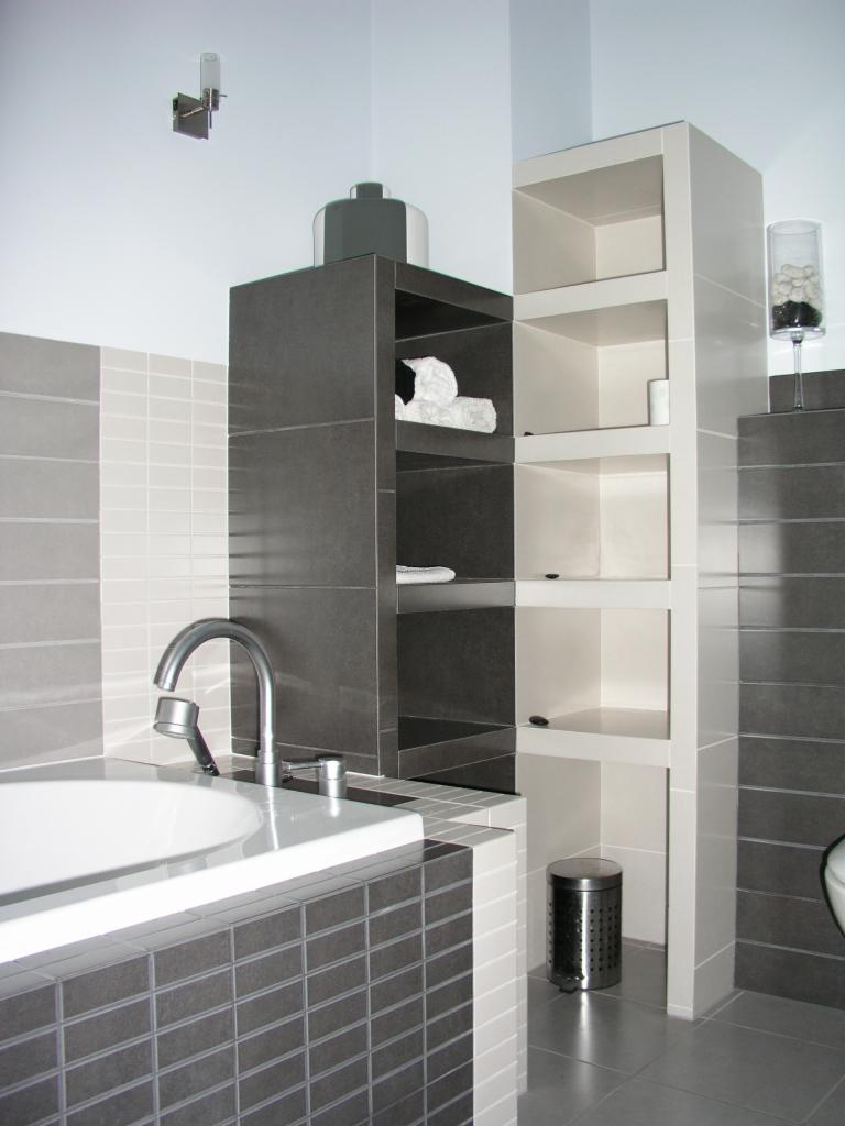 Nowoczesna łazienka z szaro-białymi płytkami, białą wanną i minimalistycznymi półkami. Elementy dekoracyjne i chromowana armatura podkreślają elegancję wnętrza.