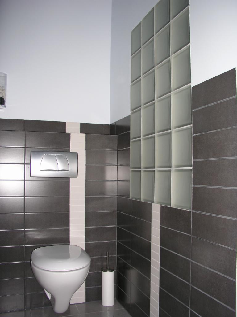 Nowoczesna łazienka z szarymi płytkami, białymi akcentami i ścianką z luksferów. Widoczna toaleta i szczotka do WC. Minimalistyczny design.