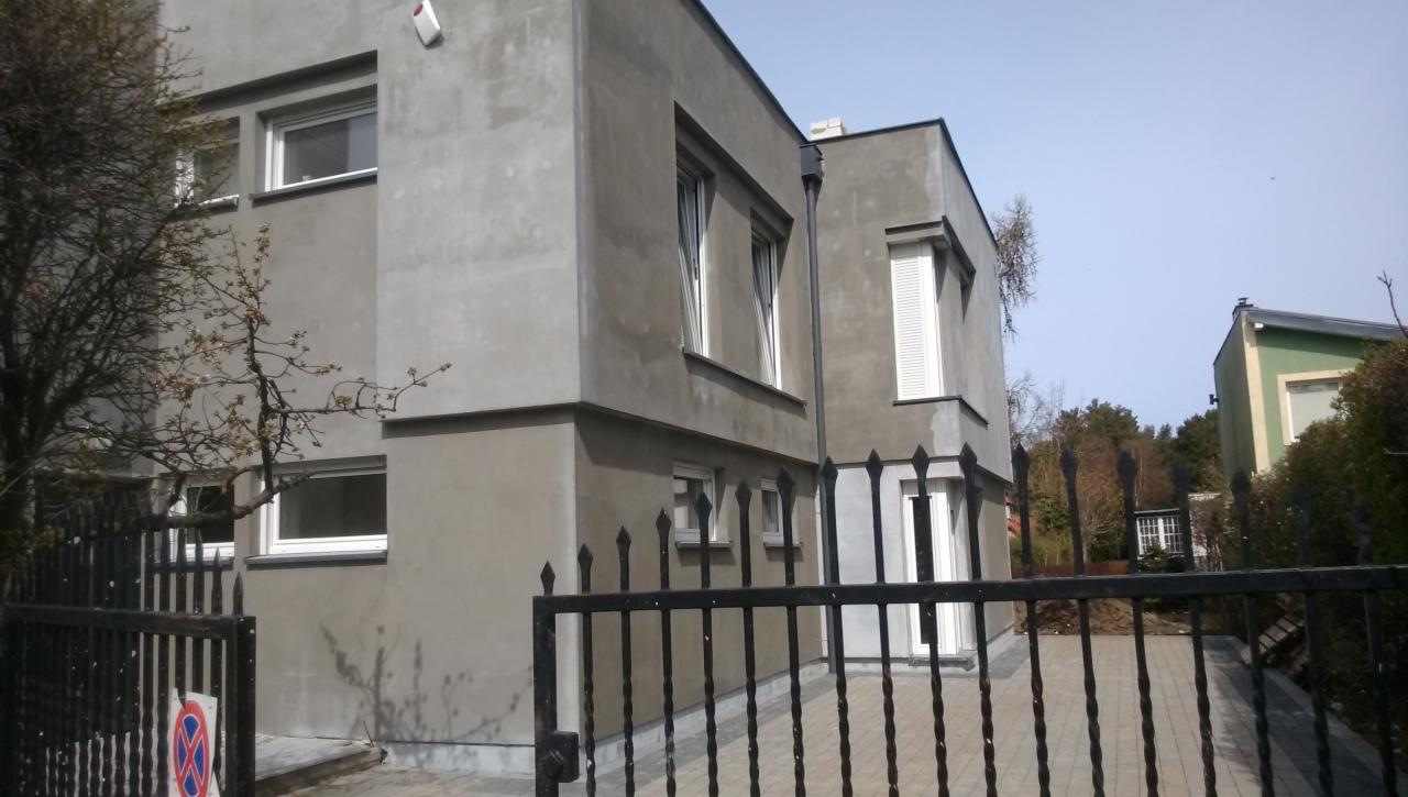 Szary, dwukondygnacyjny dom z płaskim dachem, widziany zza czarnego, kutego ogrodzenia. Widoczne okna z białymi ramami i fragment zielonego budynku w tle.