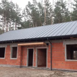 Dom w stanie surowym otwartym z zamontowaną, grafitową blachą na dachu i czarnymi rynnami, na tle lasu sosnowego.