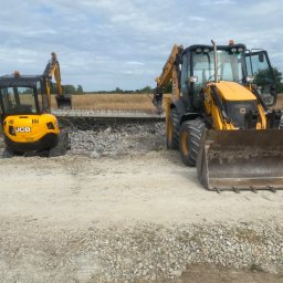 Mexi - Żółte koparki JCB na terenie budowy, gruz i zdemontowana konstrukcja w tle, krajobraz z polami pod pochmurnym niebem.