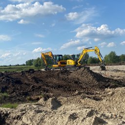Mexi - Dwa żółte mini-koparki na placu budowy z hałdami ziemi i piasku, błękitne niebo z chmurami w tle. Widoczne drzewa i trawa w oddali.