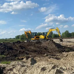 Dwa żółte mini-koparki na placu budowy z hałdami ziemi i piasku, błękitne niebo z chmurami w tle. Widoczne drzewa i trawa w oddali.