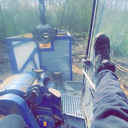 Mexi - Wnętrze kabiny maszyny do wycinki drzew z widokiem na las i mechanizm chwytający drewno. Widoczne nogi operatora w roboczych butach i spodniach.