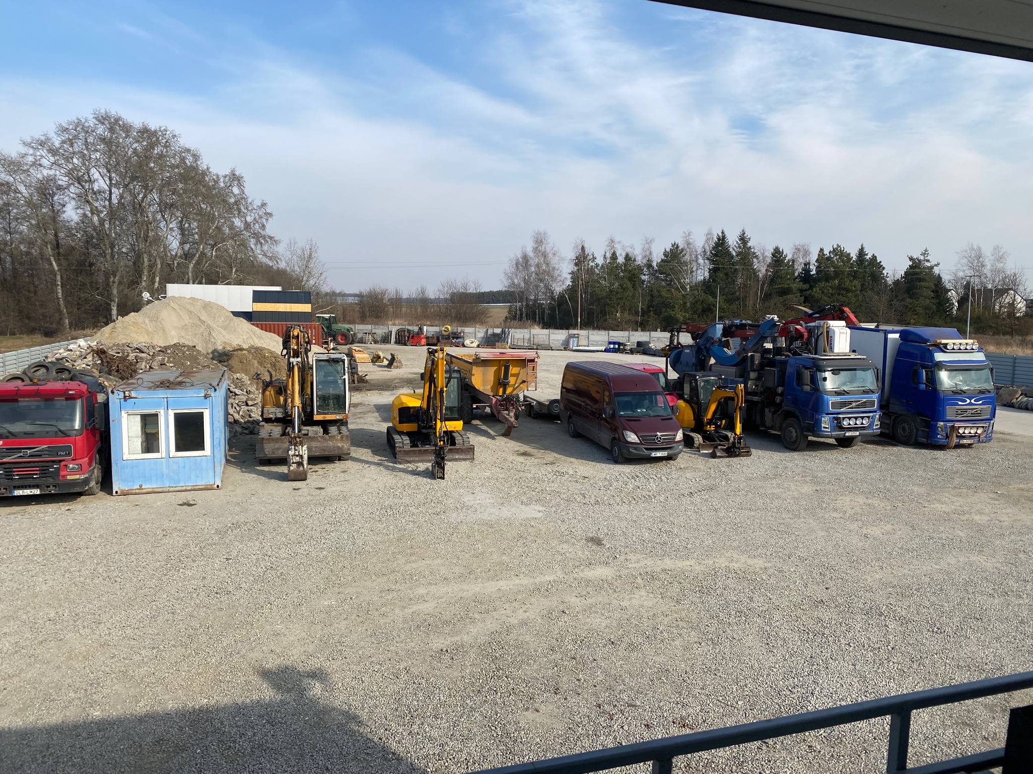 Plac z maszynami budowlanymi: koparki, ciężarówki, kontener biurowy, gruz i piasek. Widok z góry, w tle drzewa i błękitne niebo.