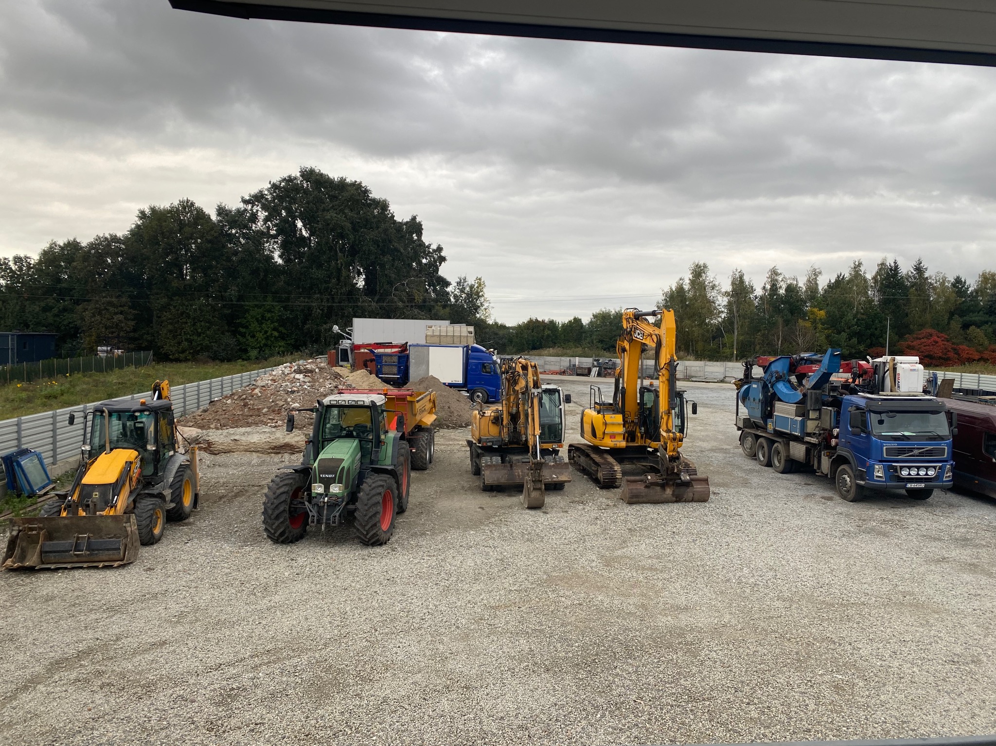 Flota maszyn budowlanych: koparki, traktory i wywrotki na żwirowym placu pod pochmurnym niebem. Sprzęt gotowy do pracy, widok z góry.