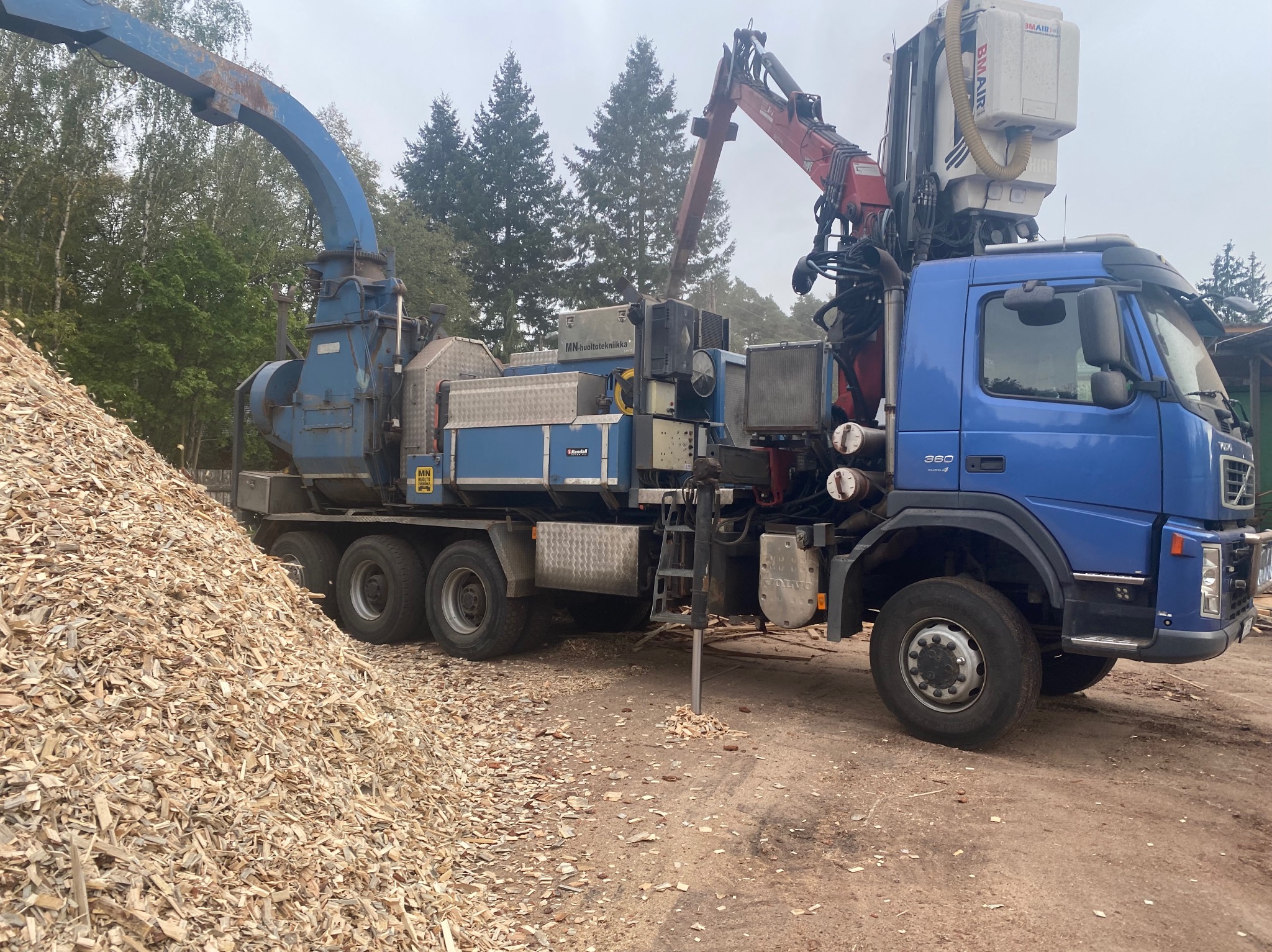 Ciężarówka z rębakiem do gałęzi i stertą zrębków na ziemi. Widoczne elementy maszyny i fragment lasu w tle. Realizacja usługi wycinki i utylizacji.