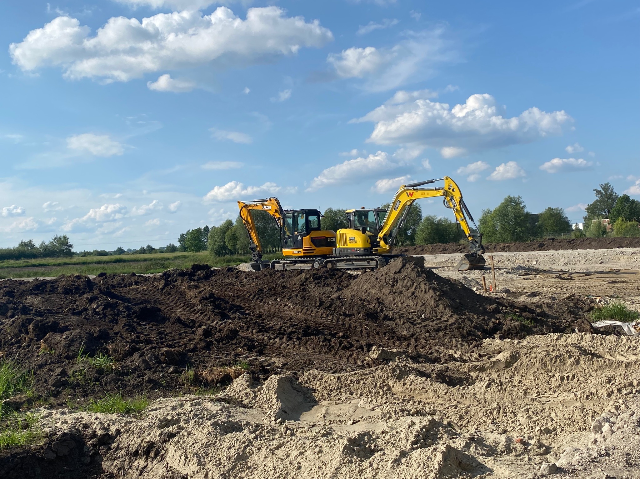 Dwa żółte mini-koparki JCB i Wacker Neuson na placu budowy, na tle ułożonej ziemi i błękitnego nieba z chmurami. Widoczne ślady opon na ziemi.