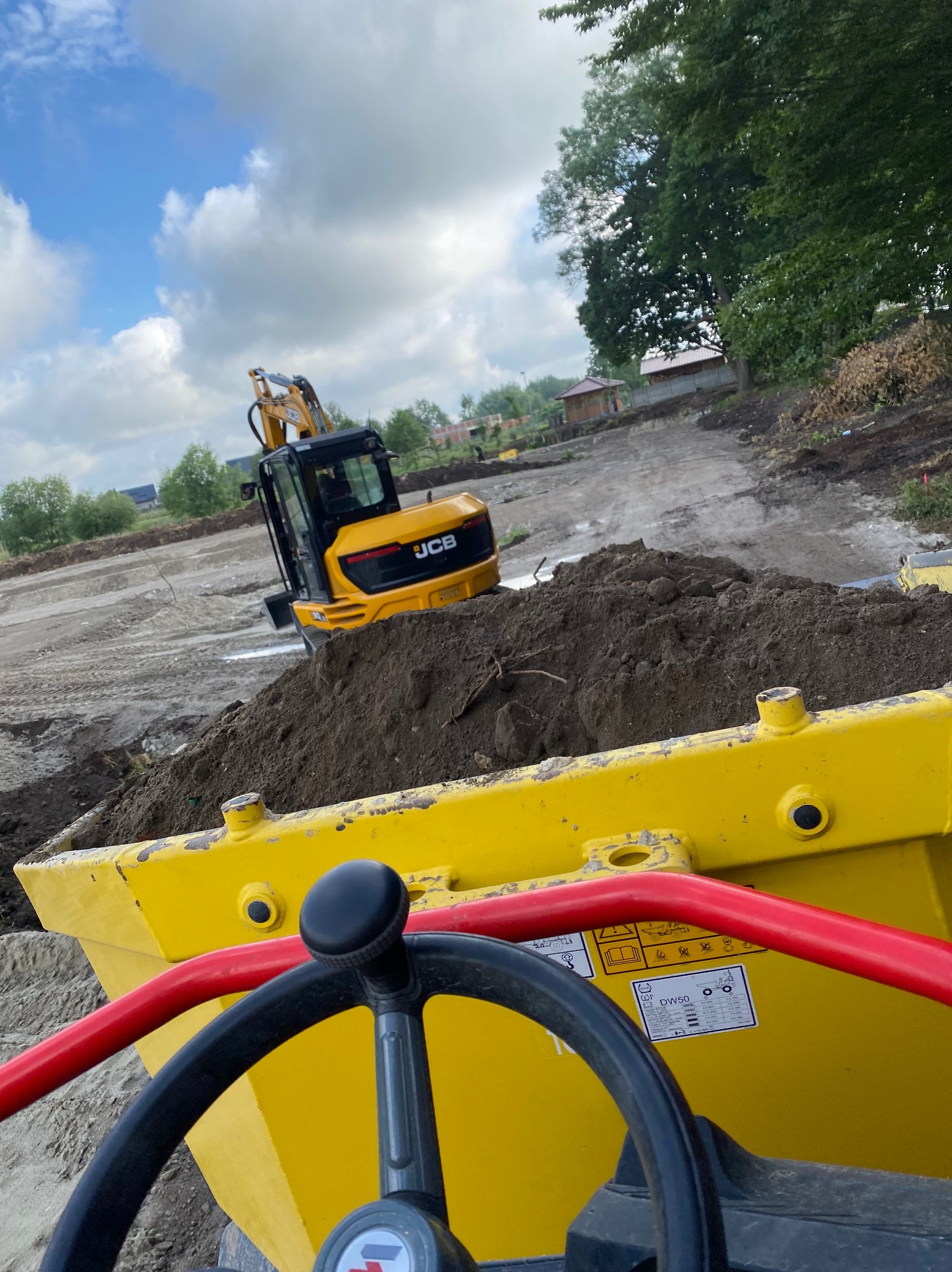 Żółta koparka JCB na placu budowy, widok z perspektywy operatora. W tle wyrównany teren i drzewa pod błękitnym niebem z chmurami.