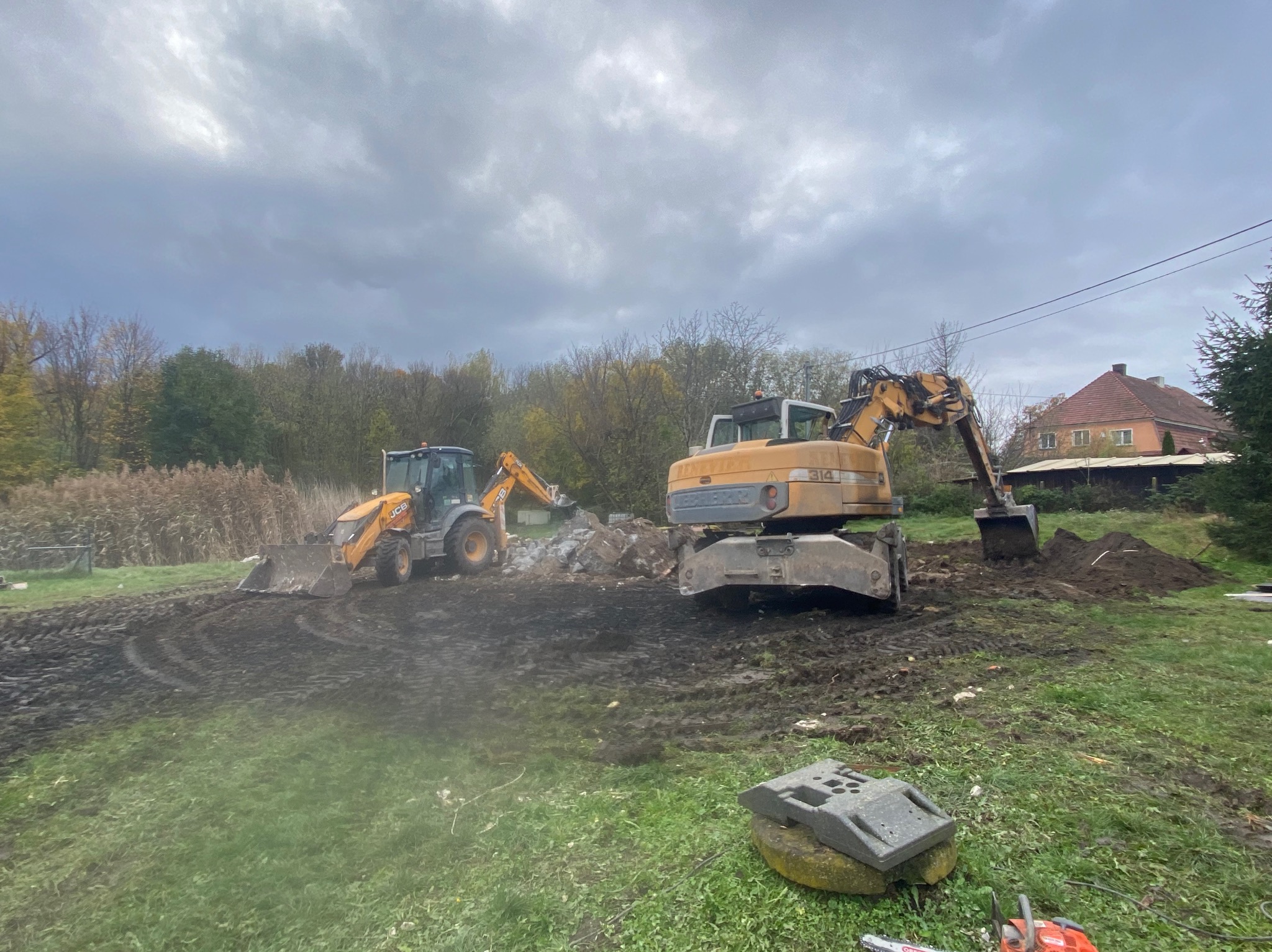 Dwie koparki pracują na błotnistym terenie. Jedna z koparek ma łyżkę zanurzoną w ziemi. W tle drzewa i pochmurne niebo. Na pierwszym planie leży piła łańcuchowa.