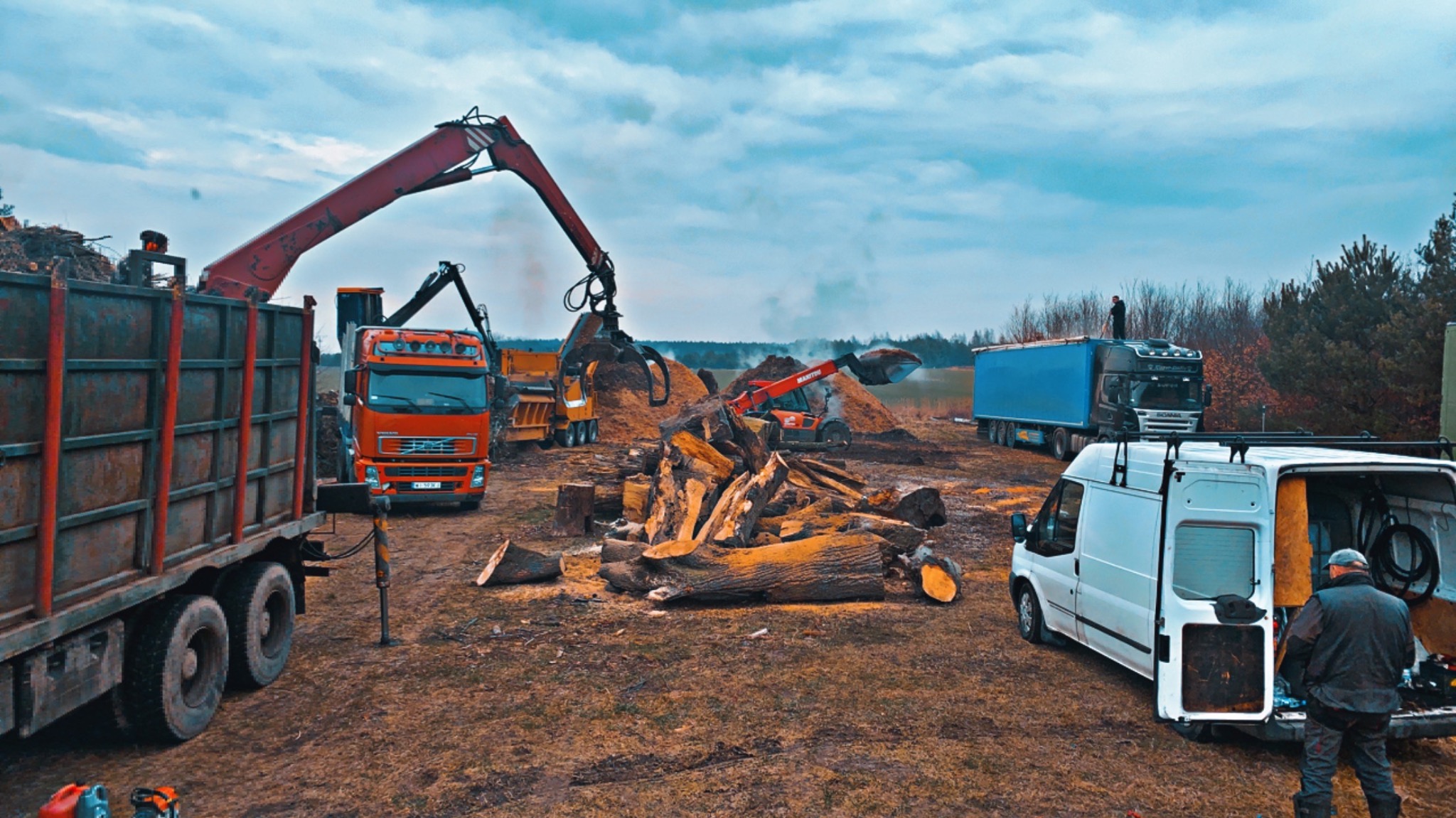 Wywóz drewna z terenu: ciężarówki z dźwigami załadowują pocięte pnie drzew. Biały bus z otwartym bagażnikiem w tle. Praca wre.