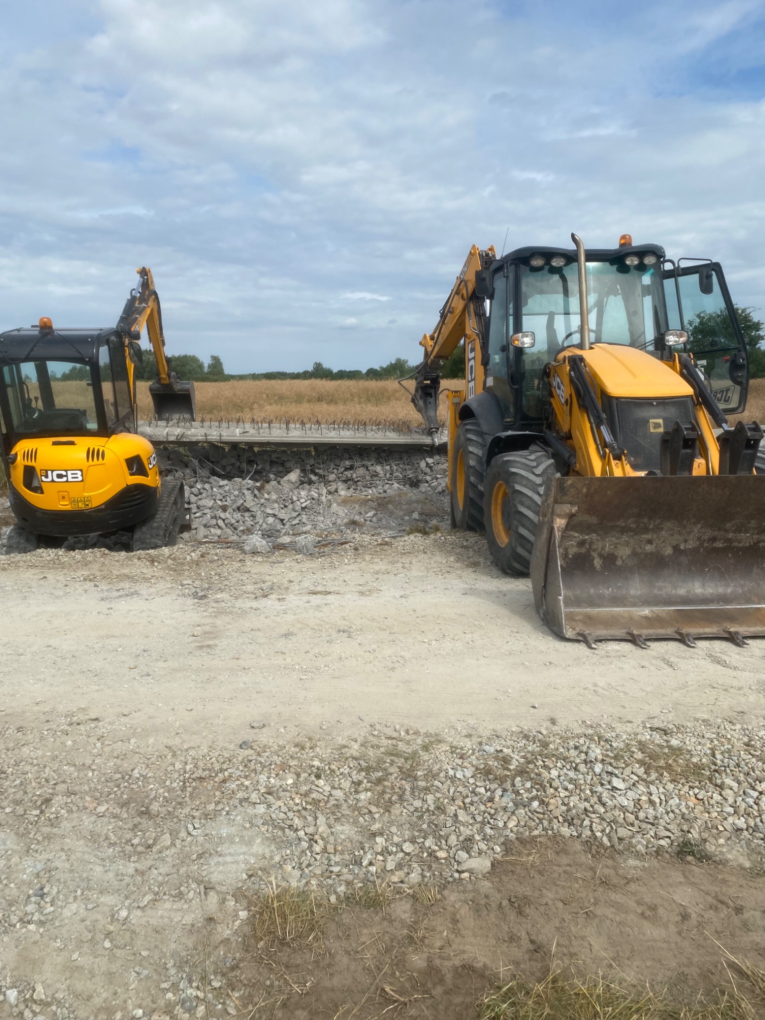 Żółte koparki JCB na terenie budowy, gruz i zdemontowana konstrukcja w tle, krajobraz z polami pod pochmurnym niebem.
