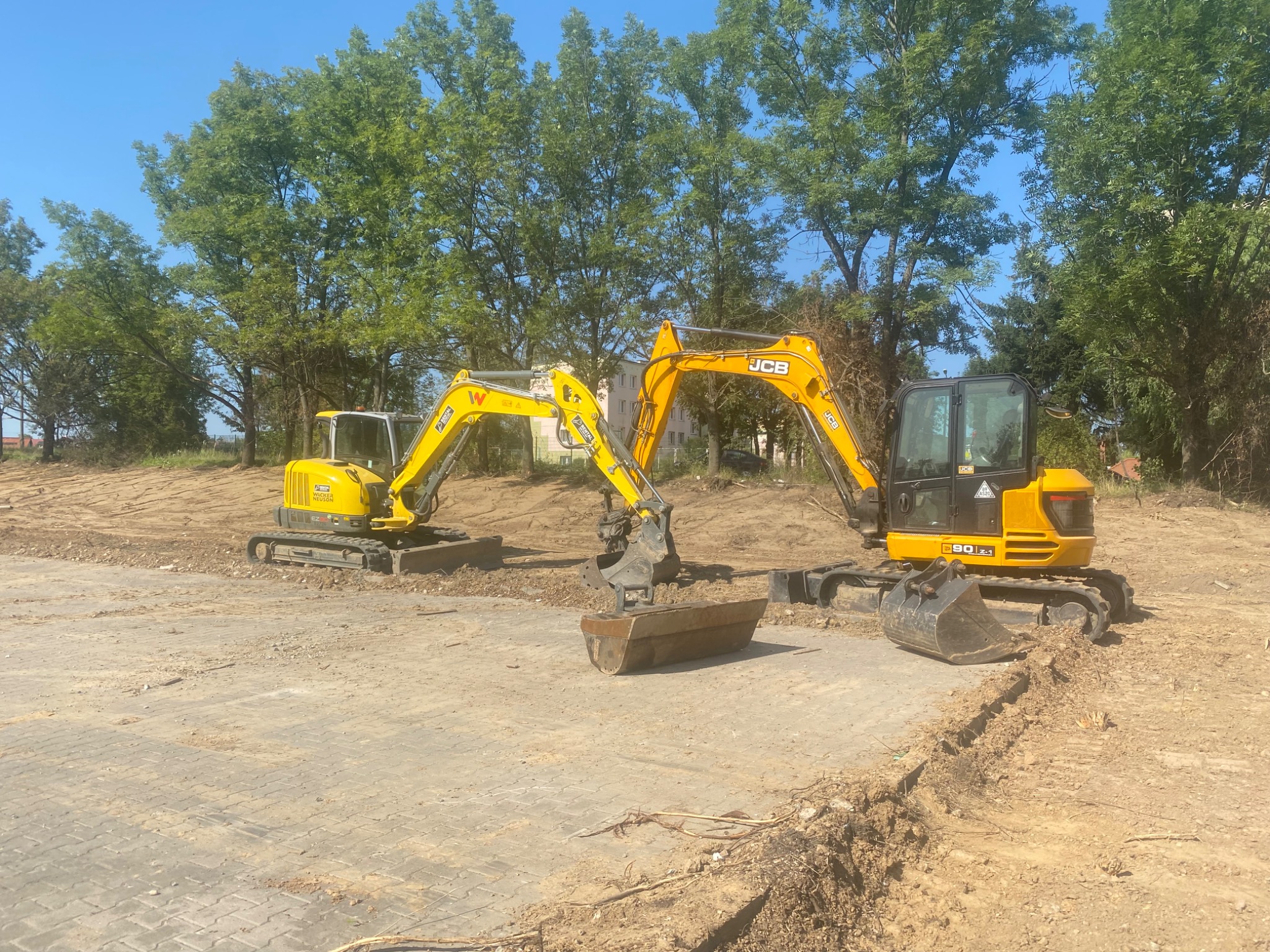 Dwie żółte koparki JCB na placu budowy, z wykopanym rowem w tle, pod błękitnym niebem i zielenią drzew. Maszyny gotowe do pracy.