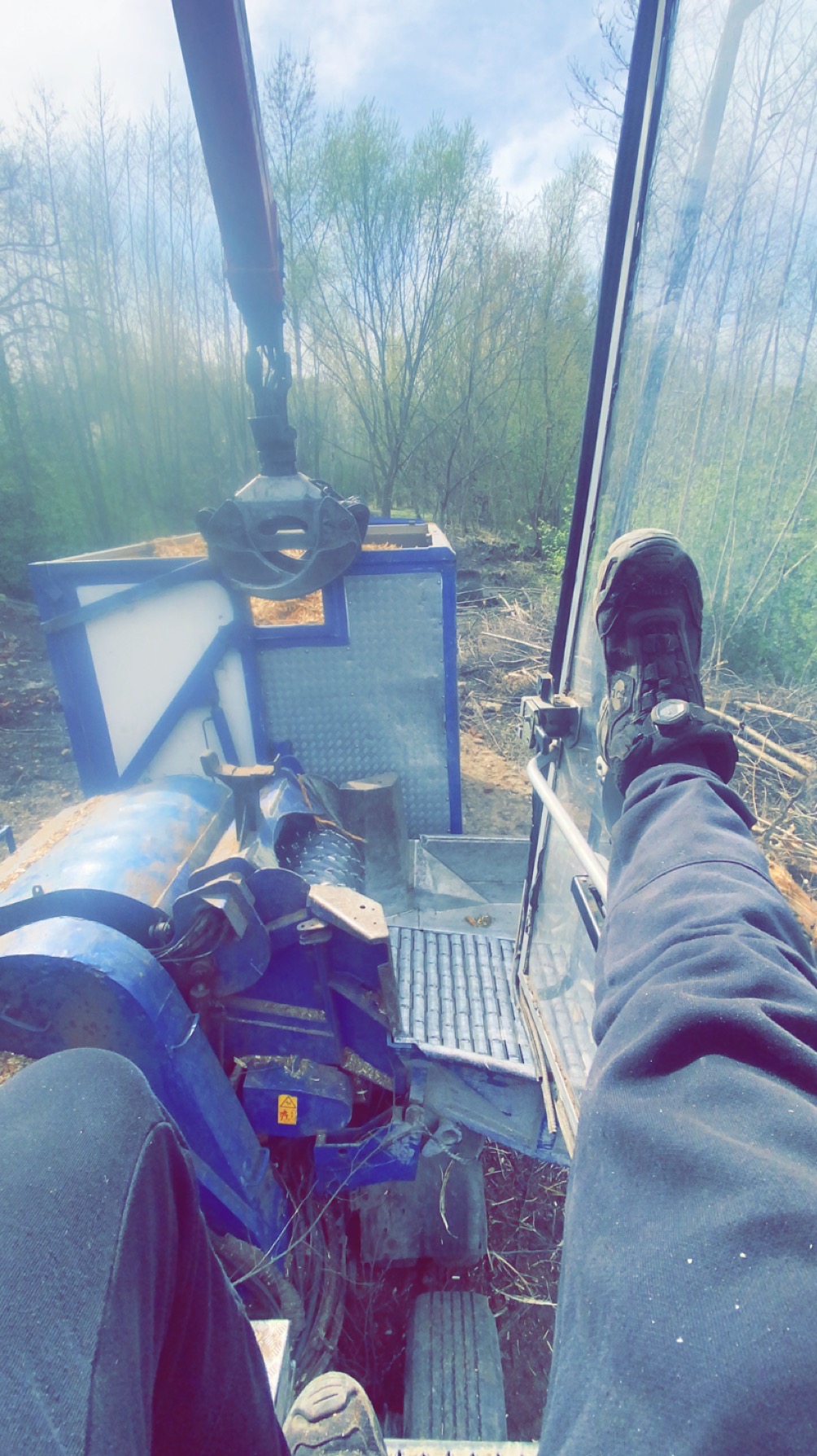 Wnętrze kabiny maszyny do wycinki drzew z widokiem na las i mechanizm chwytający drewno. Widoczne nogi operatora w roboczych butach i spodniach.