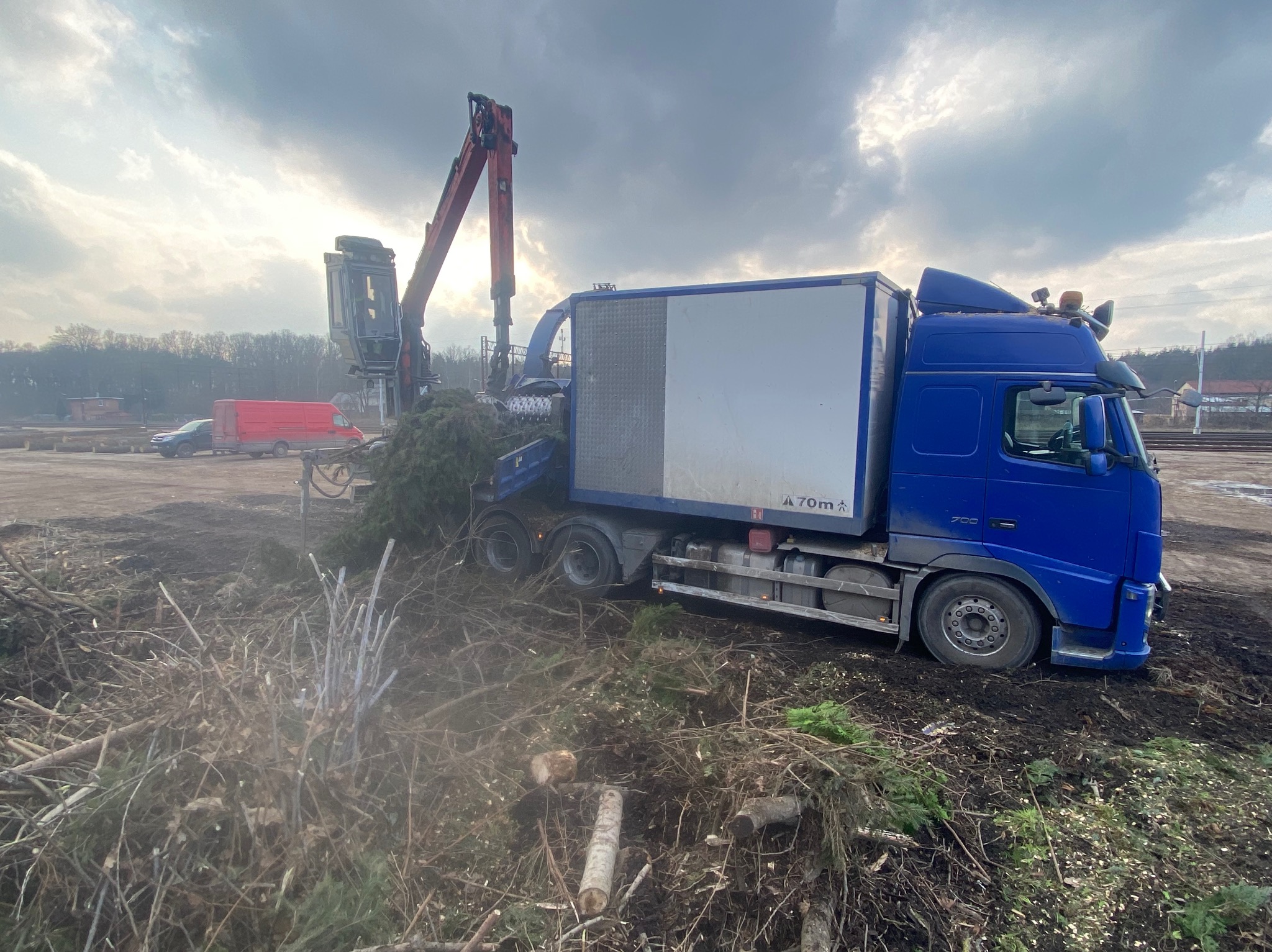 Ciężarówka z dźwigiem załadowuje gałęzie do rozdrabniacza na tle pochmurnego nieba. Widoczne ścięte drzewa i gałęzie. Praca w toku.