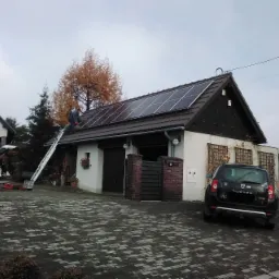 Montaż paneli słonecznych na dachu budynku gospodarczego w Jasienicy, widok z poziomu brukowanego podjazdu, drabina oparta o dach, pracownik w trakcie instalacji.