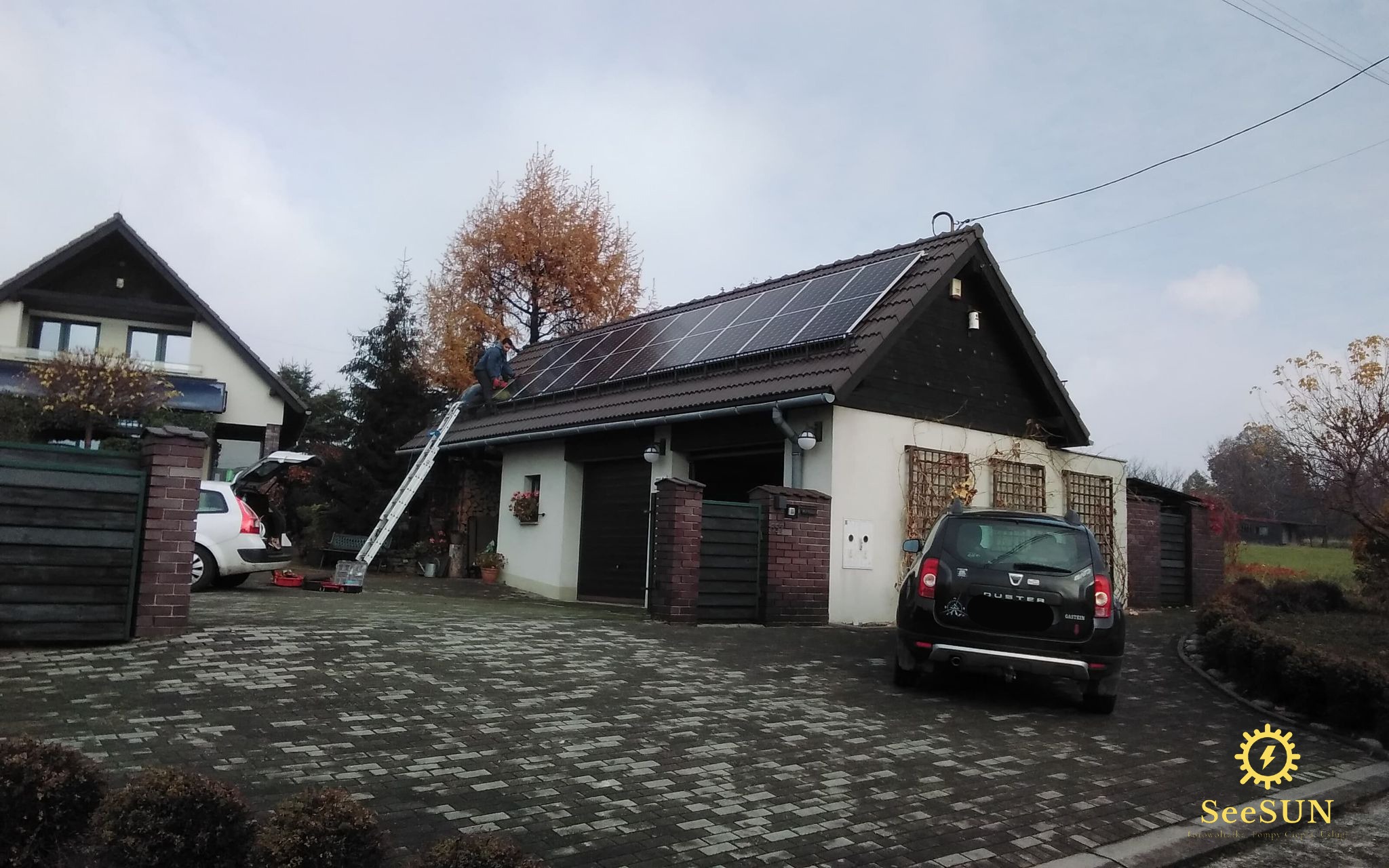 Montaż paneli słonecznych na dachu budynku gospodarczego w Jasienicy, widok z poziomu brukowanego podjazdu, drabina oparta o dach, pracownik w trakcie instalacji.