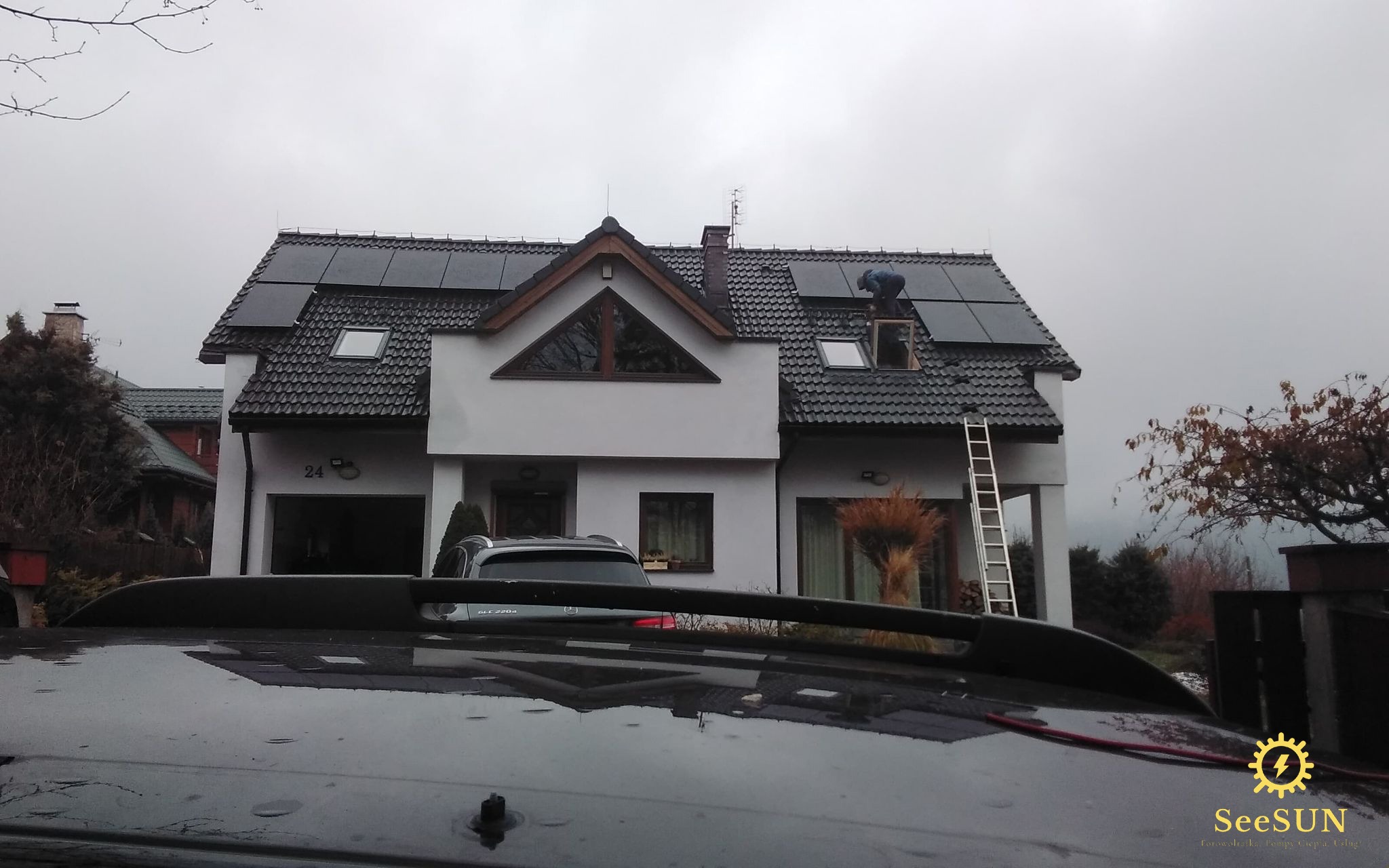 Montaż paneli fotowoltaicznych na dachu domu jednorodzinnego z drabiny, widok z perspektywy samochodu z logiem firmy SeeSUN w prawym dolnym rogu.