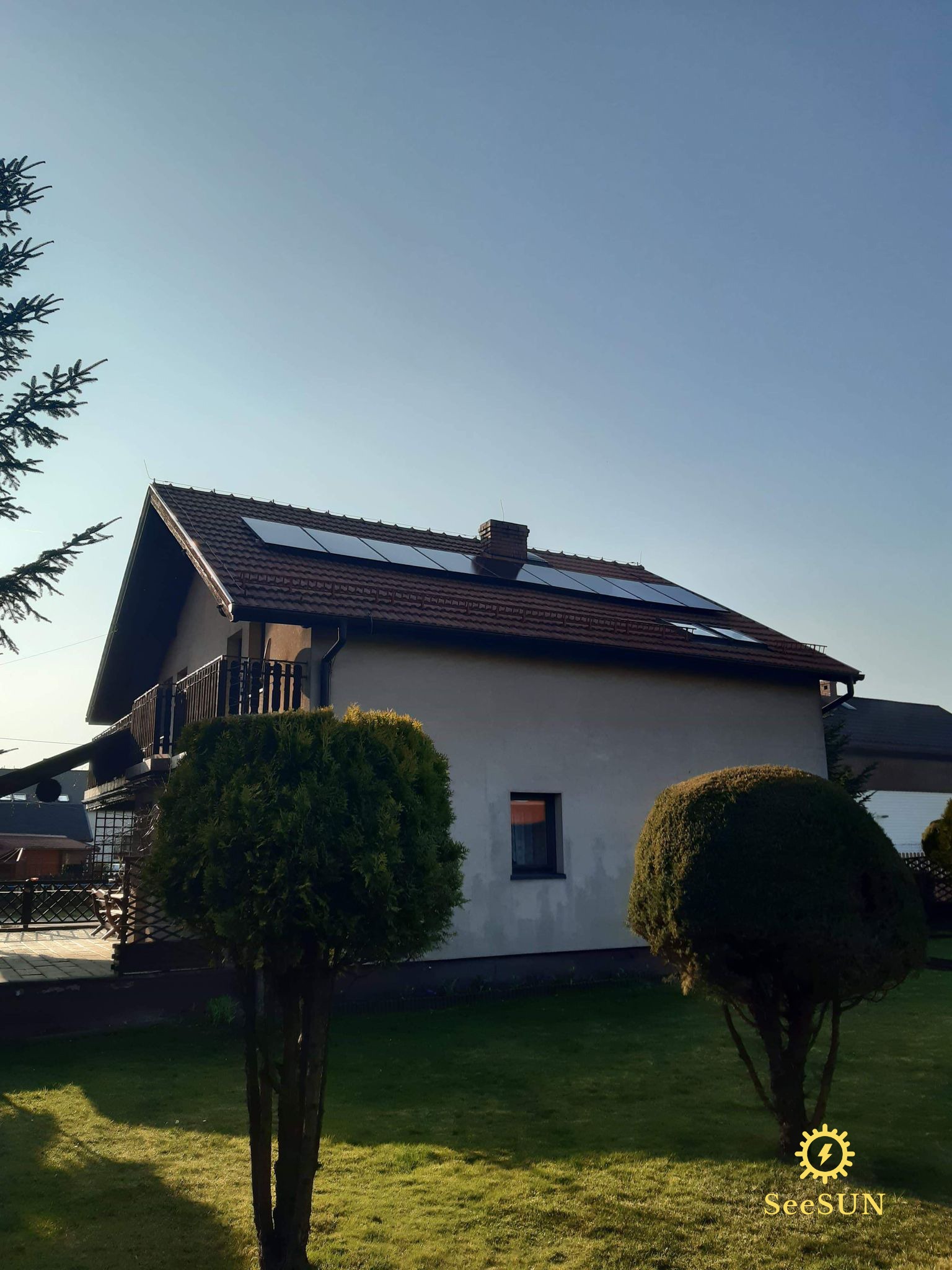 Dom jednorodzinny z zamontowanymi panelami fotowoltaicznymi na dachu, widoczne z poziomu ogrodu z dwoma kulistymi drzewami i logo firmy SeeSUN w rogu.