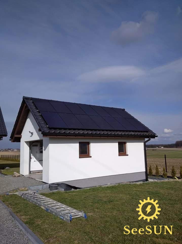 Biały budynek gospodarczy z ciemnymi panelami słonecznymi na dachu, drabina oparta o ścianę, w tle pole uprawne i błękitne niebo z chmurami.