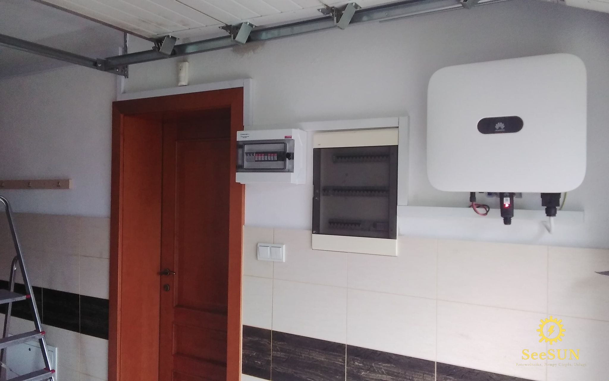 Zamontowany biały falownik marki Huawei na ścianie w garażu, obok skrzynka bezpiecznikowa i rozdzielnica, widoczna drabina aluminiowa i fragment drzwi z brązowego drewna.