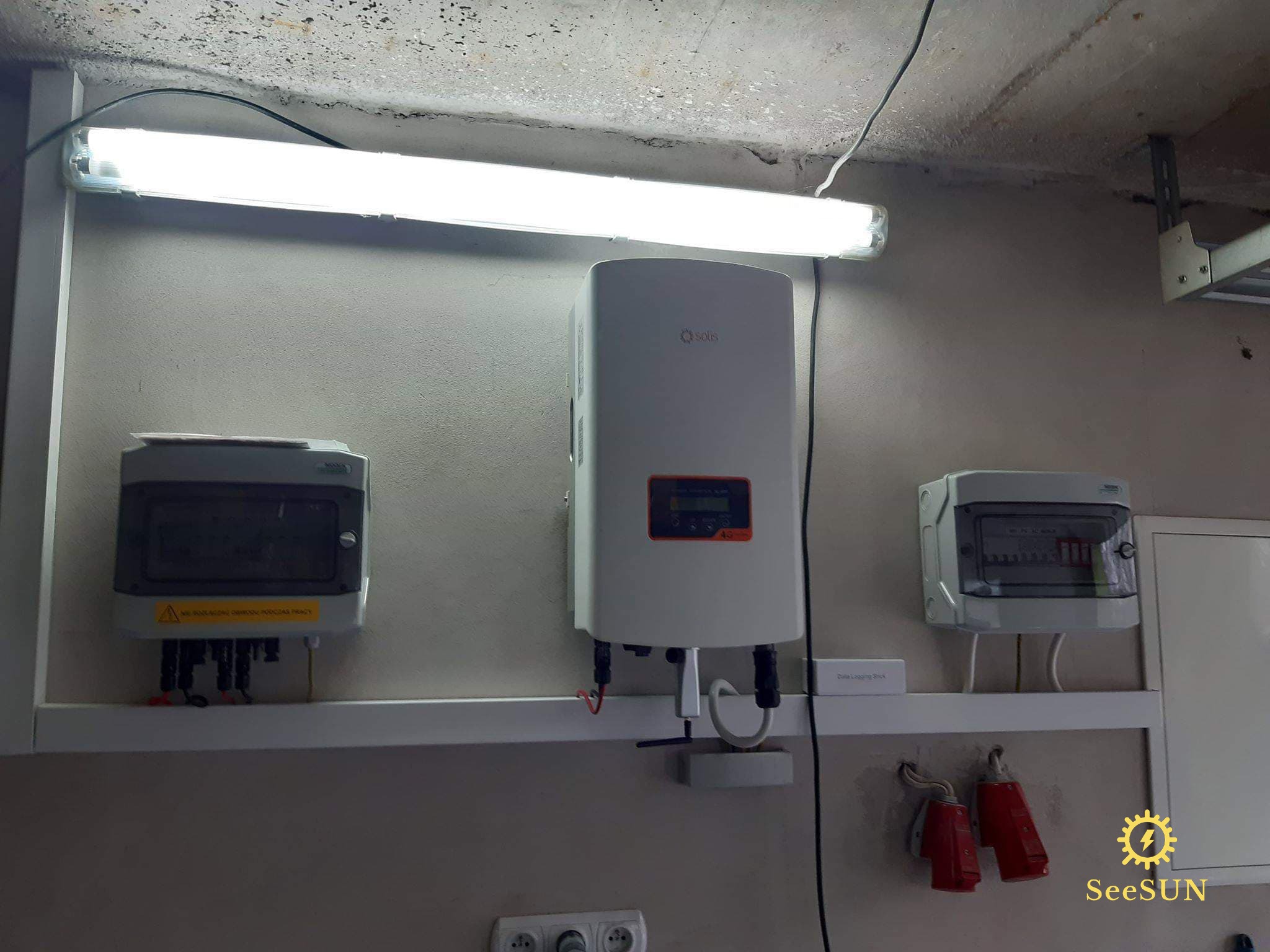 Zamontowany inwerter solarny Solis na ścianie w pomieszczeniu, obok skrzynka elektryczna i tablica rozdzielcza, oświetlone lampą jarzeniową. Widoczne gniazdka elektryczne i czerwone wtyczki siłowe.