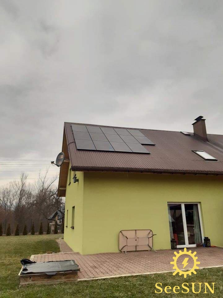 Żółty dom z brązowym dachem, na którym zamontowano panele słoneczne. Widoczne satelita, komin, okno dachowe i logo firmy na dole zdjęcia.