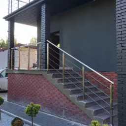 Zewnętrzne schody z ceglanym podestem i nowoczesną, poziomą balustradą ze stali nierdzewnej z elementami w kolorze złotym, prowadzące do wejścia budynku o ciemnej elewacji.