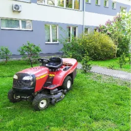Czerwona kosiarka traktorek na trawniku przed budynkiem z szarą elewacją i licznymi oknami, częściowo skoszona trawa.