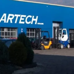Błękitny budynek z logo firmy ARTECH i rzędem wózków widłowych różnych kolorów zaparkowanych przed nim.