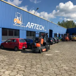 Pomarańczowy wózek widłowy Toyota na tle niebieskiego budynku firmy ARTECH z logo wózka widłowego i zaparkowanymi samochodami.