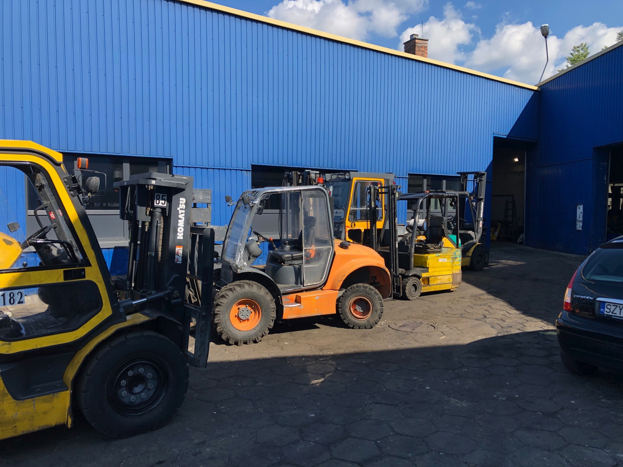 Rząd wózków widłowych różnych marek i kolorów ustawionych przed niebieskim budynkiem magazynowym w słoneczny dzień.