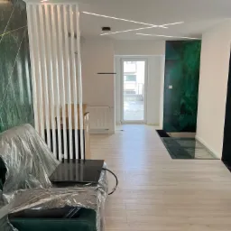 Wnętrze mieszkania w Brzostku po remoncie, z zieloną ścianą w geometryczne wzory, drewnianą podłogą i szklaną wstawką w podłodze, sofa zabezpieczona folią ochronną.